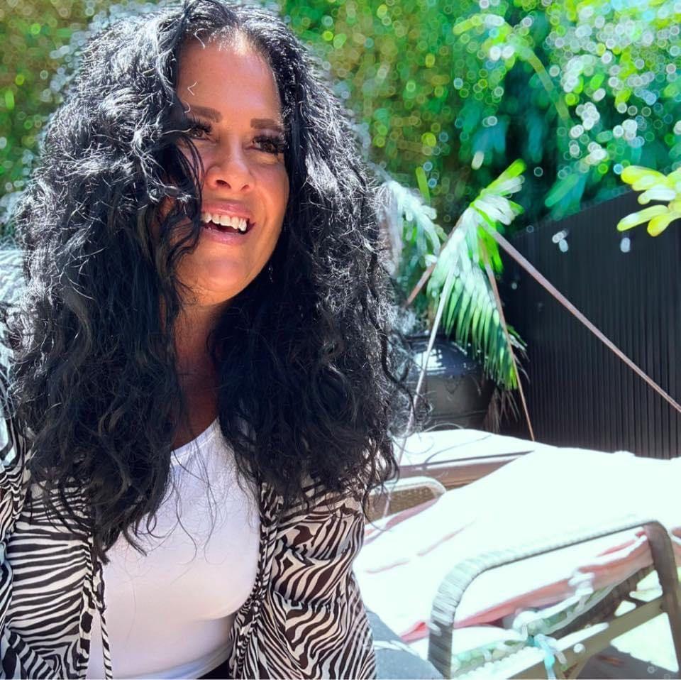 Woman with curly hair smiling outdoors with greenery and lounge chairs in the background.